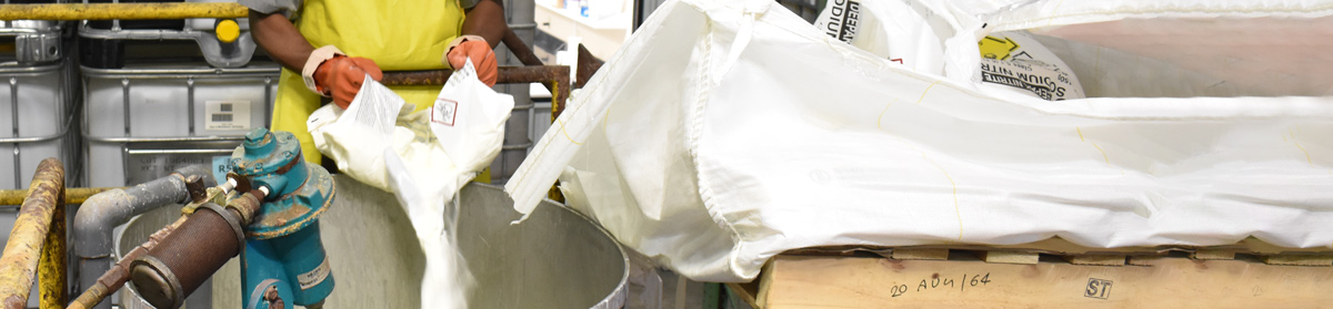 A worker wearing yellow gloves is pouring a white powder from a bag into a large metal container in an industrial or laboratory setting, with additional bags and equipment in the vicinity.