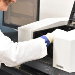 A person in a lab coat and gloves is operating a large analytical laboratory instrument, possibly inserting a sample for analysis.
