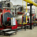 an interior view of a boiler room with several large red and silver boilers connected to yellow pipes and various gauges and valves. The industrial setup indicates a facility that likely handles significant heating requirements.