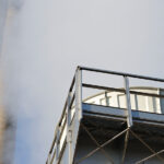a large industrial chimney emitting white smoke against a clear sky, with a metal structure featuring stairs and a platform in the foreground.