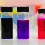 Three small vials containing blue, purple, and red liquids, with a colorful and blurred laboratory background.