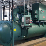 A large green industrial chiller unit with various pipes and gauges is installed inside a facility, featuring a digital control panel on the side.