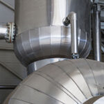 industrial ductwork and a large, spherical steel vessel, part of a mechanical system, with various pipes connected to it inside a facility.
