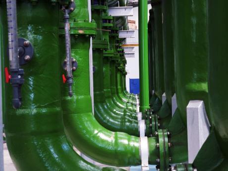 Large green industrial pipes with valves and supports run along a wall, part of a complex machinery system.