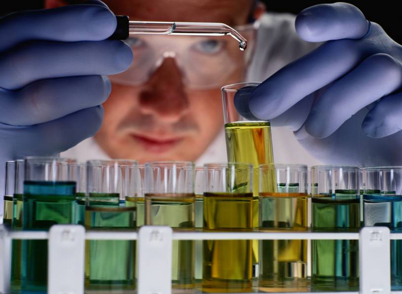 A scientist in safety goggles and gloves is pipetting a liquid into a test tube, with rows of test tubes containing green and yellow liquids in the foreground.