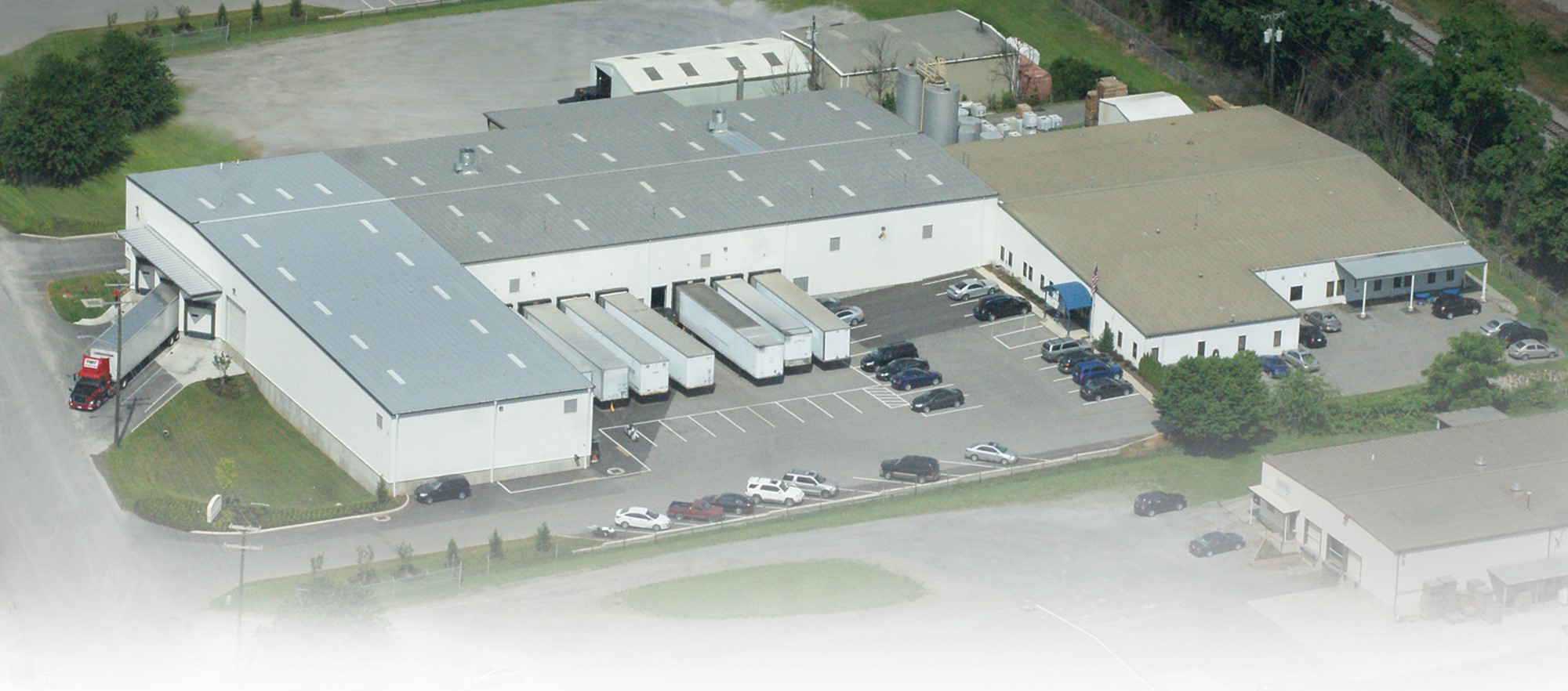 An aerial view of a large industrial building complex with a parking lot, several docked delivery trucks, and adjacent smaller structures.