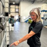 Woman with gray/blonde hair wears a black QualiChem polo and a silver wristwatch points at something on a QualiChem warehouse shelf.